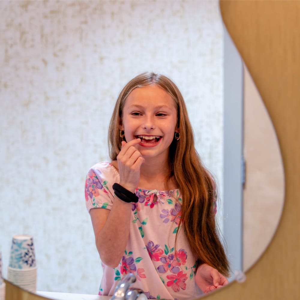 A young girl smiles and looks at her teeth in the mirror.
