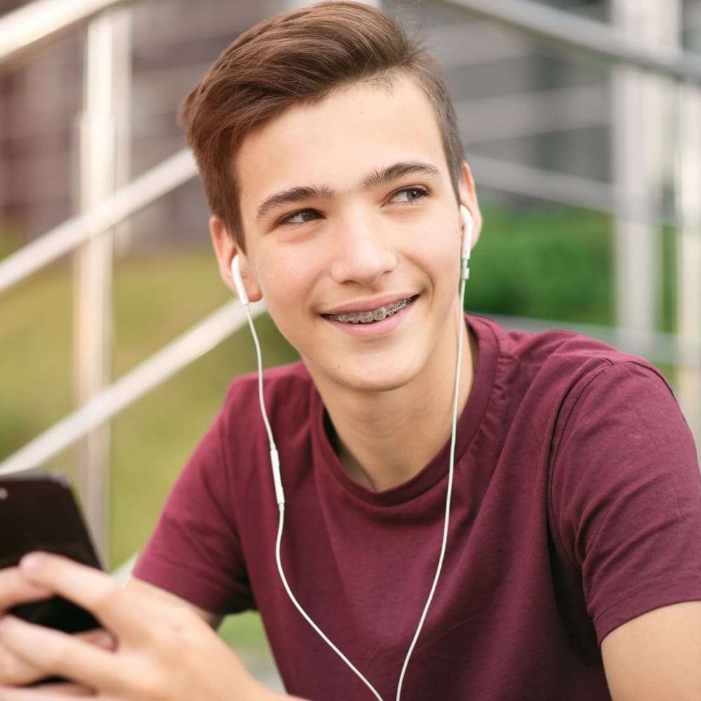 A teenage boy is listening to headphones and smiling. His smile reveals he has braces.